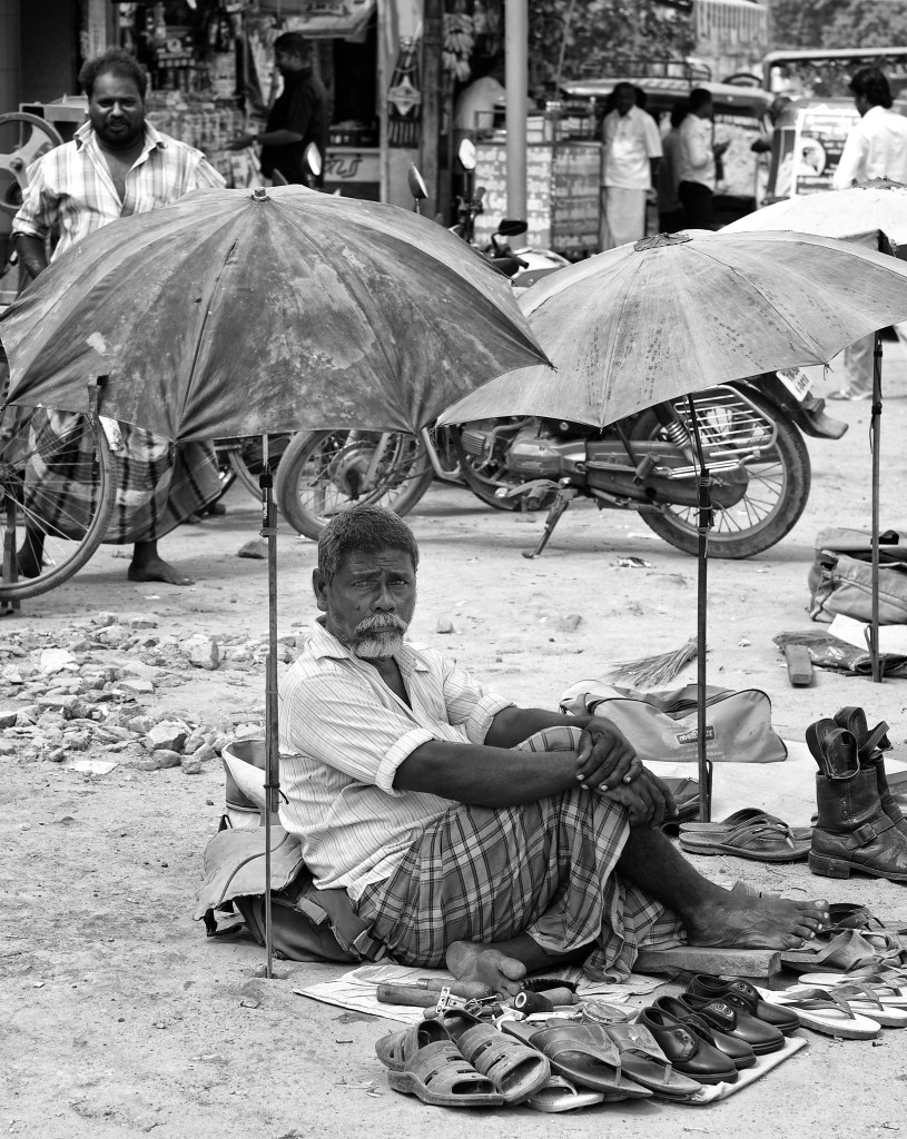 A shoemaker setup his workplace by roadside with an umbrella for shade from the Sun, Virudhunagar, India