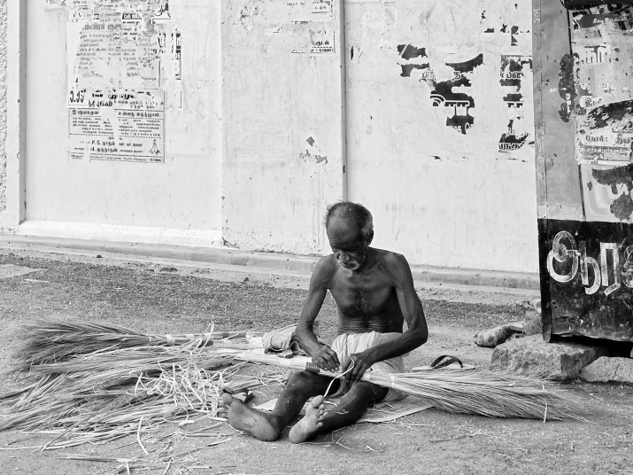 An old man tying bunch of bristles and making brooms for sale by the roadside. Virudhunagar, India. 
